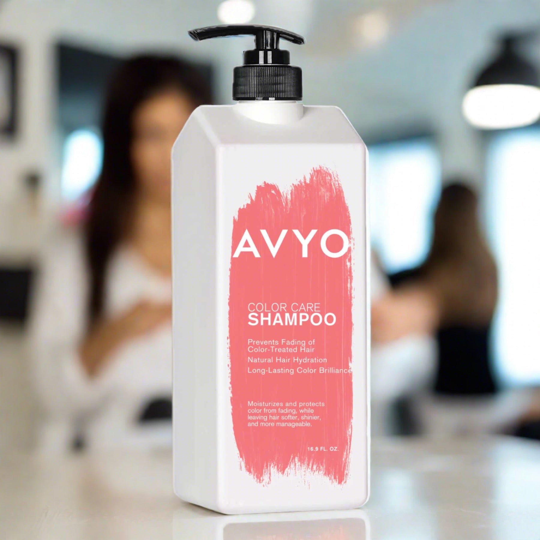 A bottle of AVYO Color Care Shampoo is prominently displayed on a counter with a blurry background featuring a woman in a salon. The white bottle has a bold red brush stroke design, with text that highlights its benefits: prevents fading of color-treated hair, ensures natural hair hydration, and promises long-lasting color brilliance. It contains 16.9 FL OZ and mentions that it moisturizes, protects from fading, and leaves hair softer and more manageable.