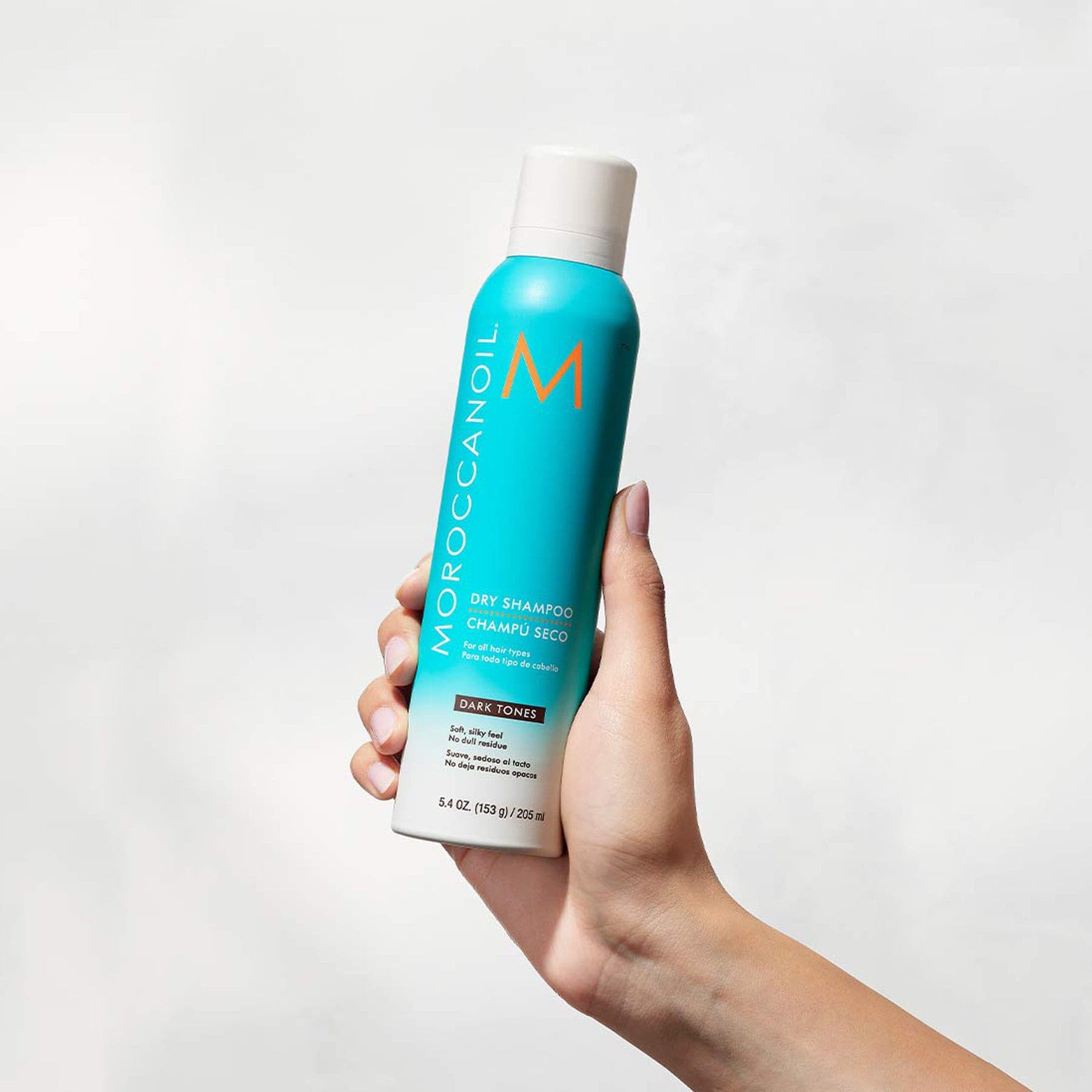 A woman right-hand holds up a Moroccanoil Dry Shampoo Dark Tones, 5.4 Fluid Ounces / 205 Milliliters.